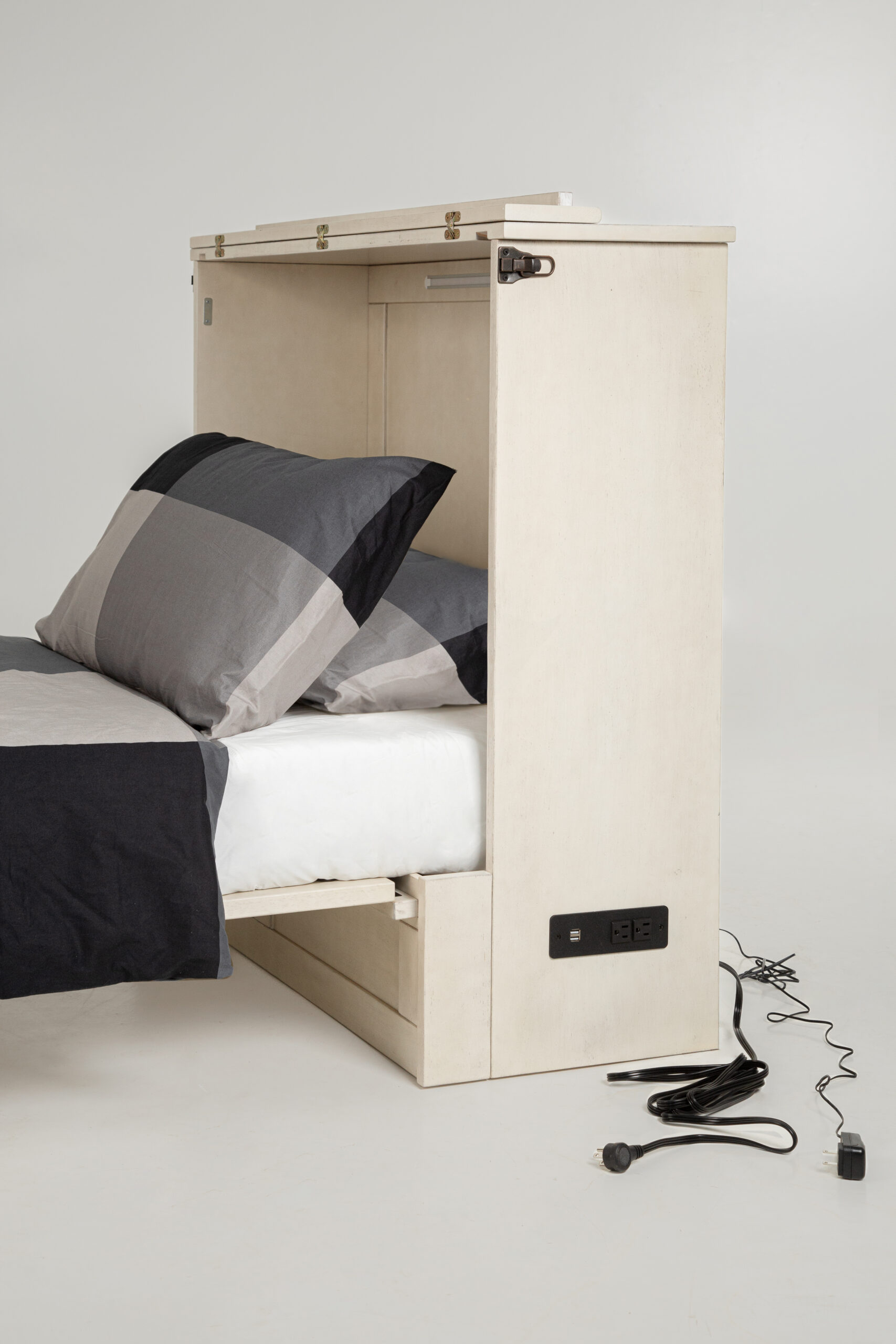 Murphy Express Twin Provence Side View - SDS Cabinet Beds Bed with a tall, light wood headboard/side panel and plaid gray‑black bedding; several cords and a power strip lie on the floor nearby, emphasizing built‑in storage.